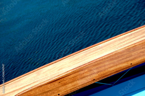 Varnished wooden side deck of a sailing boat, visible small section of light blue cockpit and large area of dark blue water overboard. photo