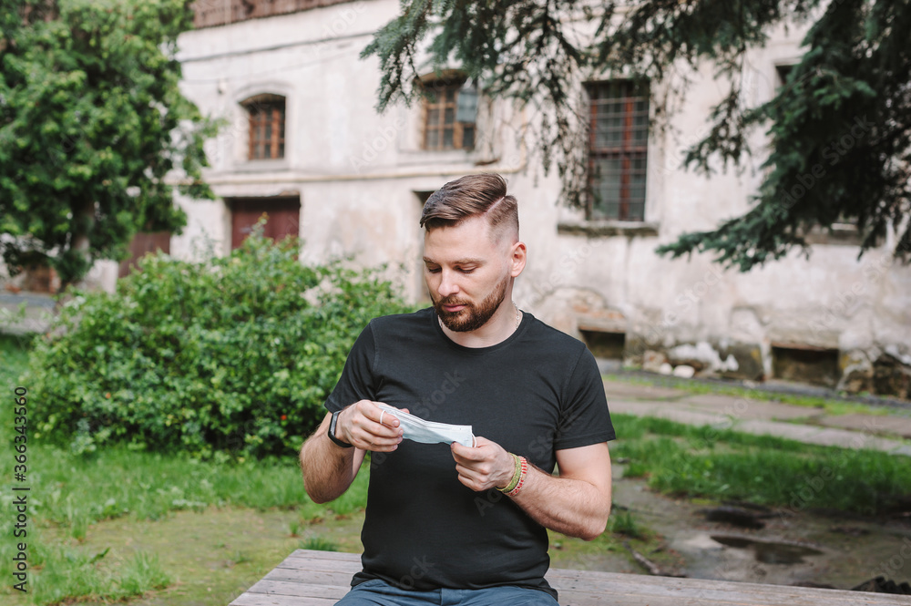 Obraz premium A bearded man holds a face mask in his hands. A man on the street is thinking whether to wear a medical disposable mask. People do not want to wear masks.