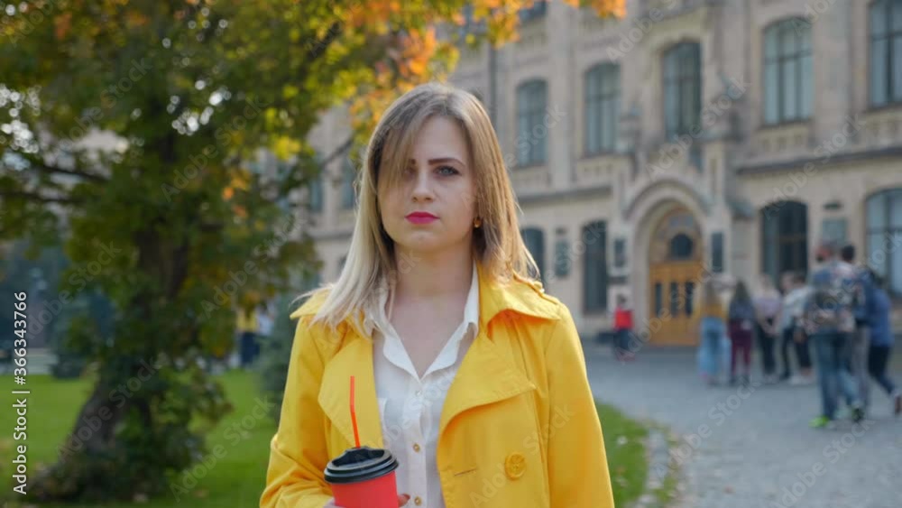 Woman in white blouse, yellow cloak with coffee in hands walks through the park. Girl with pink lipstick on lips looks around in square. 4K slow motion footage Woman in white blouse, yellow cloak with coffee in hands walks through the park. Girl with pink lipstick on lips looks around in square. 4K slow motion footage