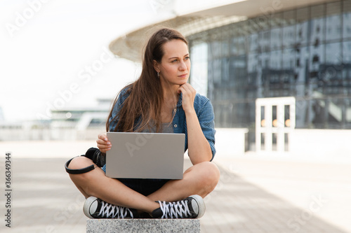 Young fashionable girl uses laptop outdoors and listens to music