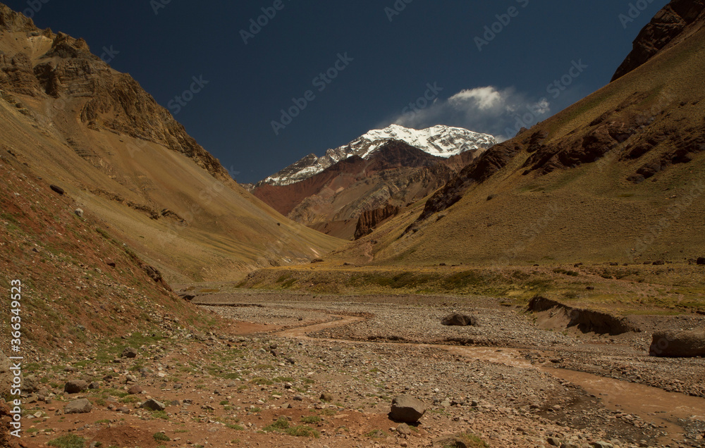 Seven summits. View of a stream flowing across the rocky and arid valley with mountain Aconcagua snowy peak in the background.