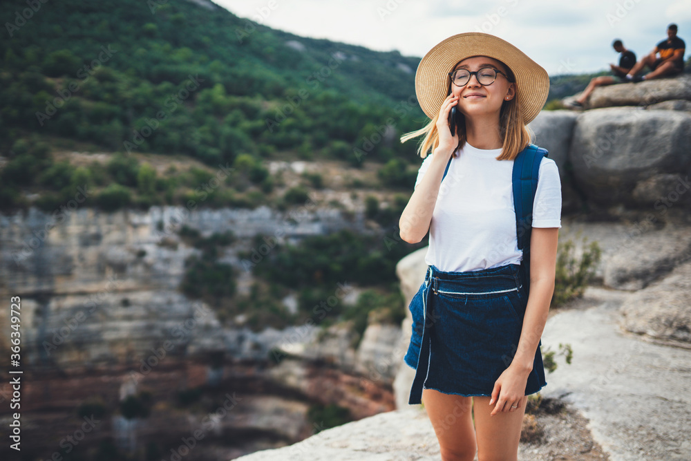 smiling girl traveler in hat and glasses communicates call on mobile phone relaxing on top of green mountain landscape, female tourist using cellphone on background of nature outside