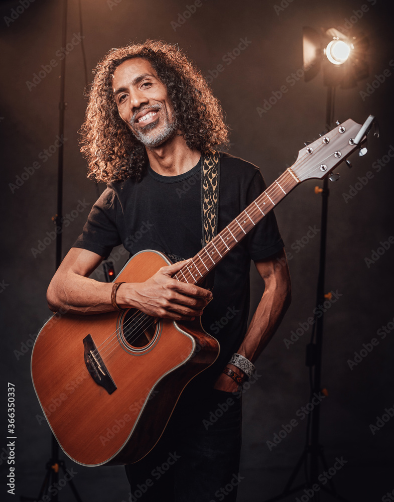 Obraz premium Middle aged hispanic man musician in black t-shirt holds guitar on a scene, smiling and looking on camera. View of musician in the spotlight