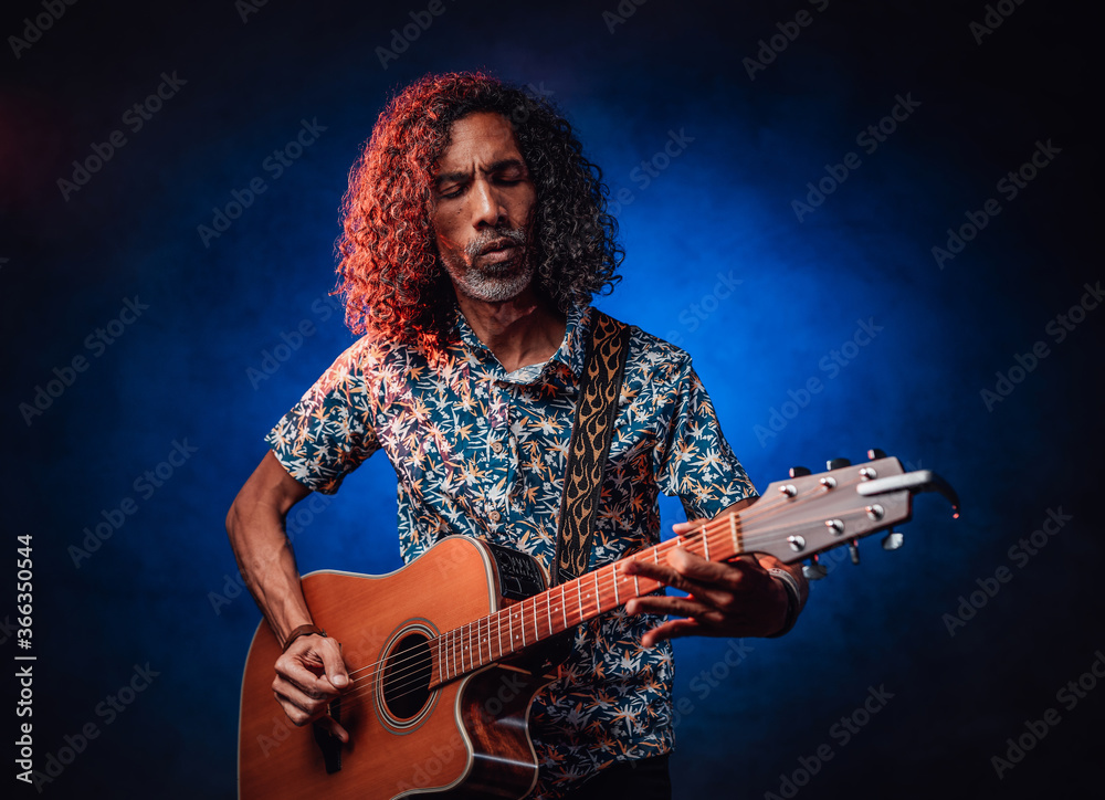 Obraz premium Talented hispanic musician in a hawaiian shirt playing guitar on a dark illuminated by blue and red light. Concept of music, hobby, festival