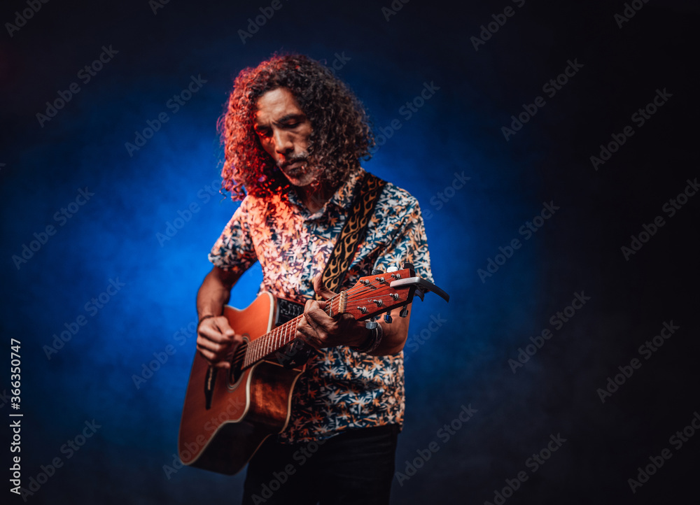 Talanted hispanic musician in a hawaiian shirt playing guitar on a dark illuminated by blue and red light. Concept of music, hobby, festival
