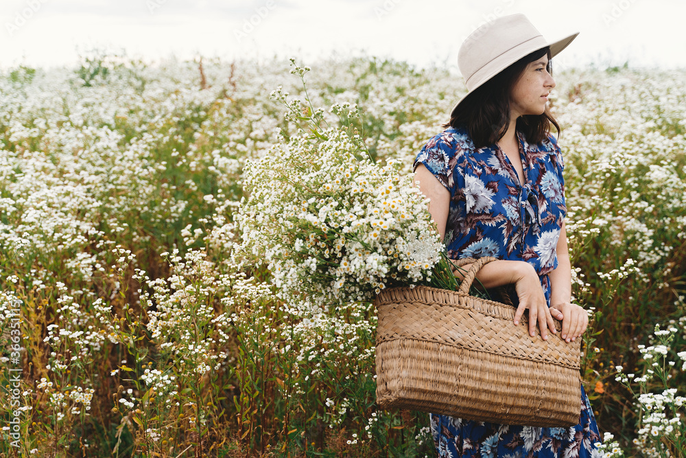 Obraz premium Stylish young woman in blue vintage dress and hat gathering white wildflowers in straw basket in meadow. Beautiful girl holding white daisies bouquet. Tranquil summer in countryside