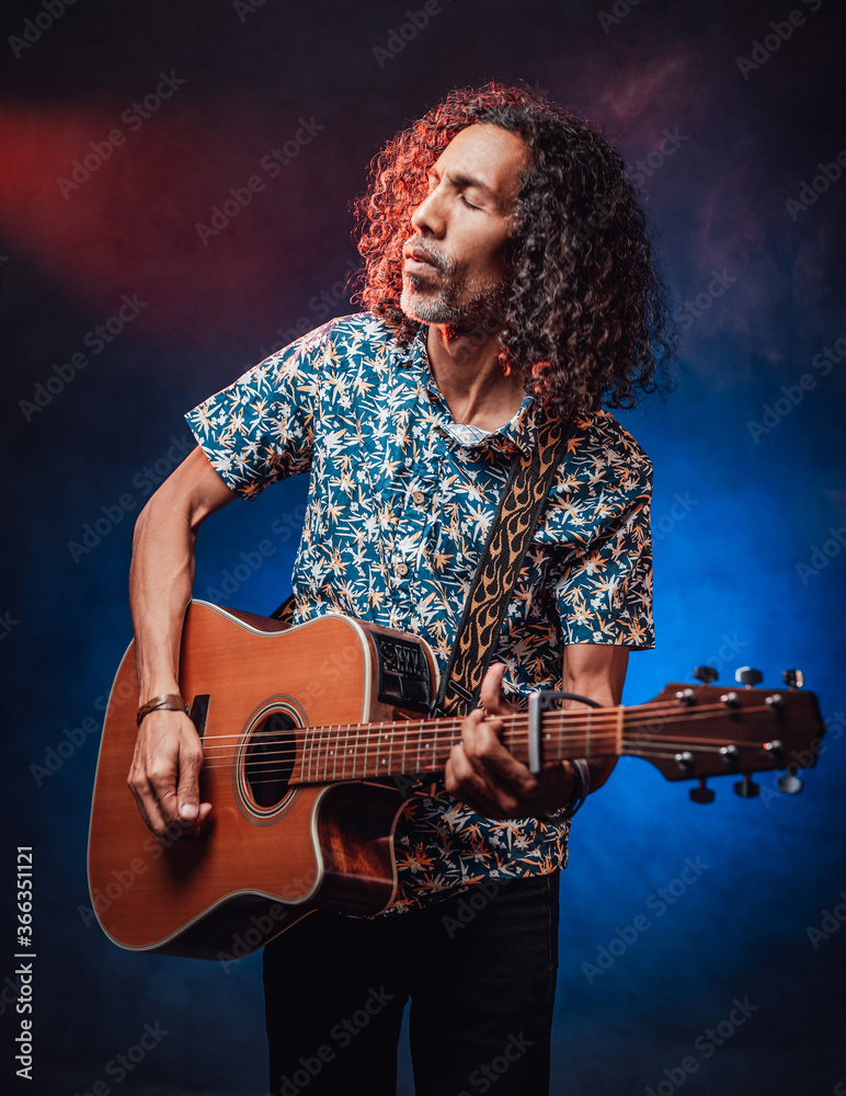 Fototapeta premium Talented hispanic musician in a hawaiian shirt playing guitar on a dark illuminated by blue and red light. Concept of music, hobby, festival