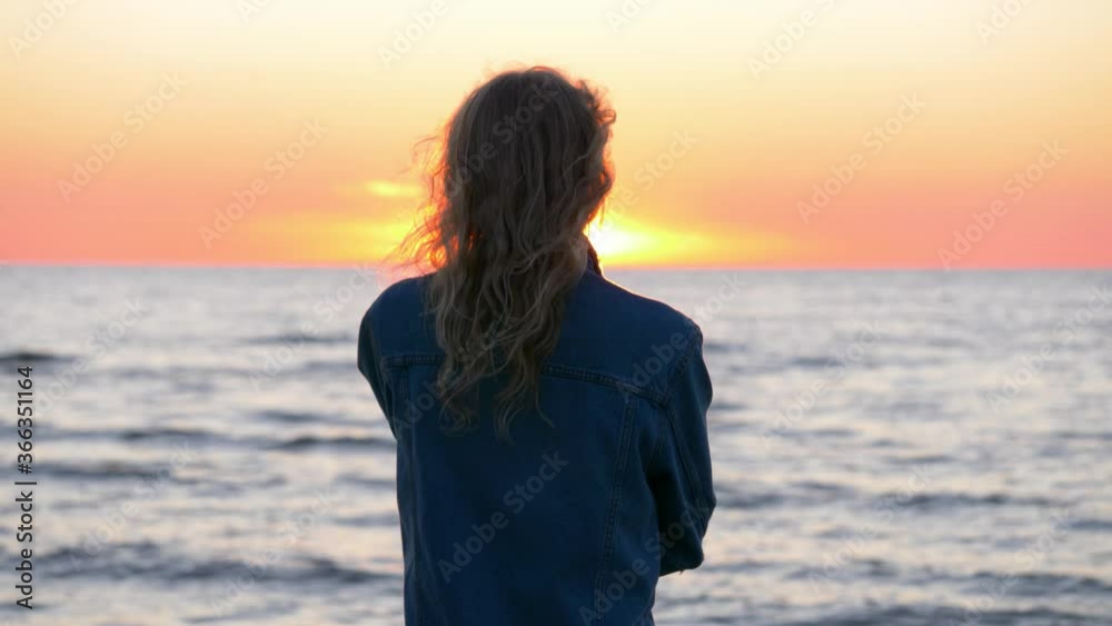 Close up shot of woman standing and watching the sunset near the sea. Beautiful blond wavy hair shakes in the wind. Point of view. Relaxed female enjoys her freedom. Close up shot of woman standing and watching the sunset near the sea. Beautiful blond wavy hair shakes in the wind. Point of view. Relaxed female enjoys her freedom.