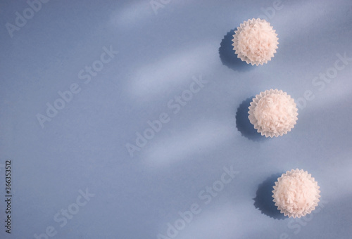 Fototapeta Naklejka Na Ścianę i Meble -  White Raffaello balls in coconut flakes in a row on blue background. Copy space. Valentine's background. Candies in a line. The sun's rays fall from the window.