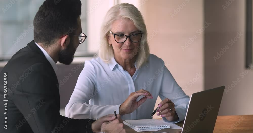 Old middle aged female professional bank manager investment advisor consulting commercial customer arabic eastern businessman explaining online project offer on laptop computer at business meeting