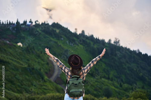 Free inspired woman tourist with backpack stands with open arms and enjoys peaceful and tranquility while traveling in the mountains