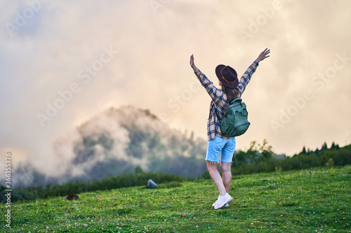 Happy free inspired woman traveler with backpack stands with open arms and enjoys calmness and tranquility while traveling in the mountains