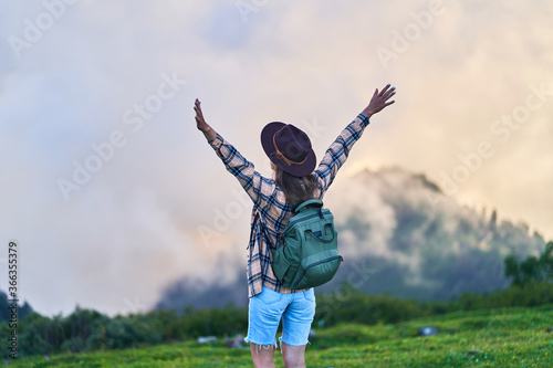 Free inspired woman traveler with backpack stands with open arms and enjoys peaceful and tranquility while traveling in the mountains