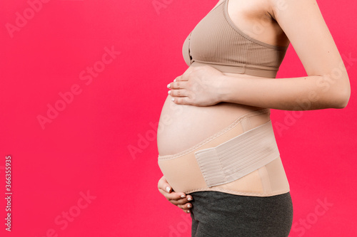 Side view of pregnant woman wearing elastic maternity band at pink background with copy space. Close up of orthopedic abdominal support belt concept © sosiukin