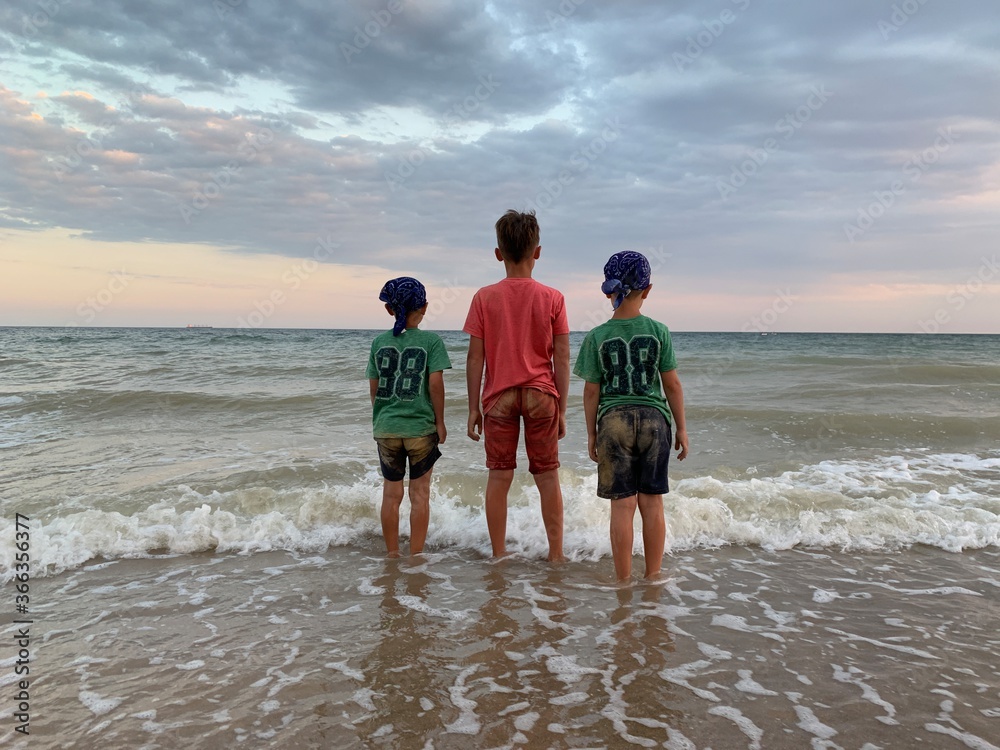 Two twins and a boy are standing by the sea. Children in the same clothes are looking at the sea. Brothers boys in hats on the beach, back view. Concept: vacation, friendship, Two twins and a boy are standing by the sea. Children in the same clothes are looking at the sea. Brothers boys in hats on the beach, back view. Concept: vacation, friendship,
