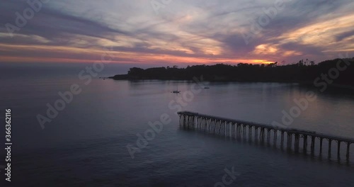 Aerial Footage of Long Pier in Bay with dark Island Forest silhouette and Beautiful Sunset with Pink Sky while camera moves further and higher away