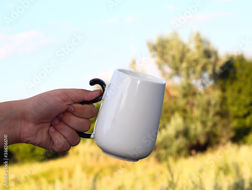 White mug in the hand of a man against the background of nature.