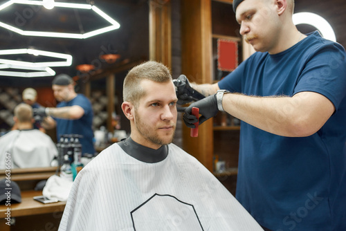 Getting new trendy haircut. Professional barber working with hair clipper, serving young man sitting in barbershop chair. Focus on a client