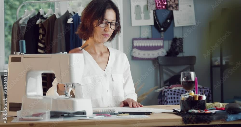 Mature female designer sitting at own fashion studio drinking red wine during creative working process. Professional workspace with sewing machine, threads, scissors and other tools for sewing. Mature female designer sitting at own fashion studio drinking red wine during creative working process. Professional workspace with sewing machine, threads, scissors and other tools for sewing.