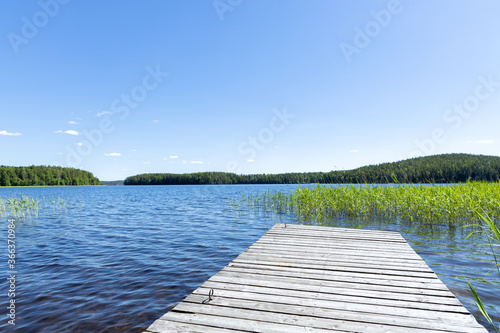 Fototapeta Naklejka Na Ścianę i Meble -  Traditional Finnish view. Beautiful calm lake on a summer morning and an old rustic wooden pier. Sun shining on forest and woods in blue sky.