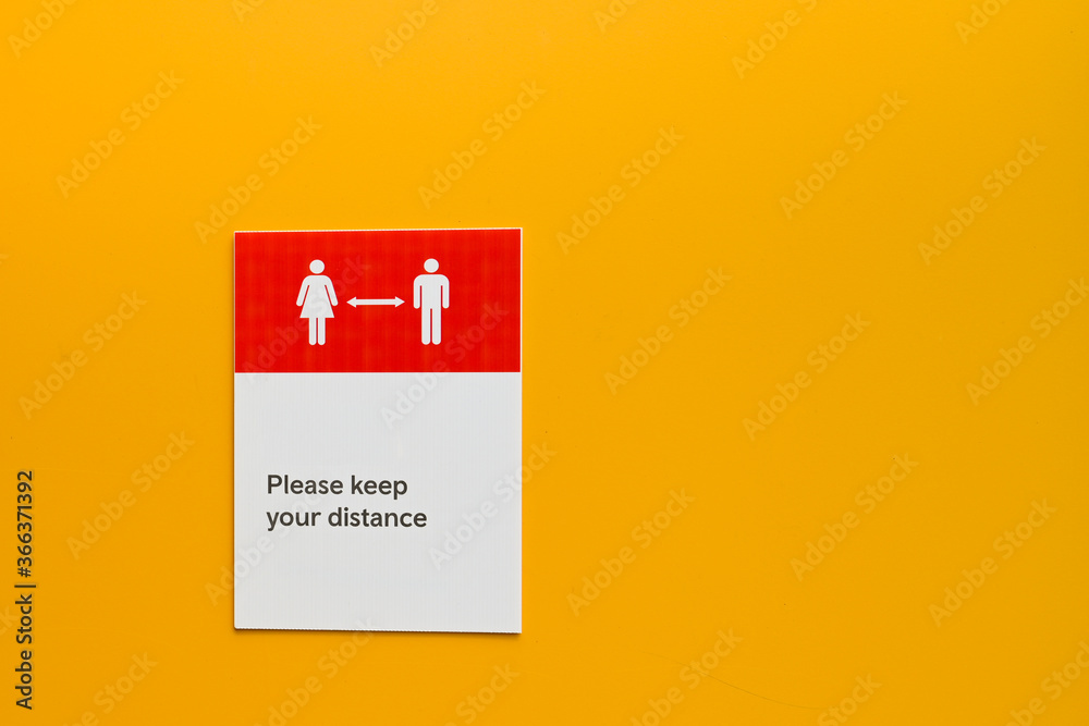 Notice on a plain yellow wall asking people to maintain social distancing. No people. Copy space. Notice on a plain yellow wall asking people to maintain social distancing. No people. Copy space.