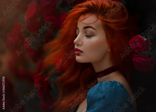 Portrait of a red haired girl in blue dress. Pretty young model between red roses, wind in the hair.Art work. photo