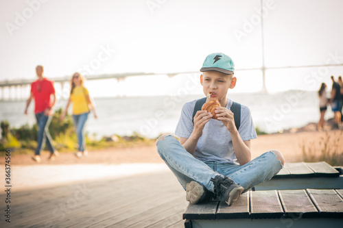The boy sits on a bench on the waterfront with his legs crossed and eats a croissant. A teenager in the street eats pastries and looks thoughtfully into the distance. People are passing by.
