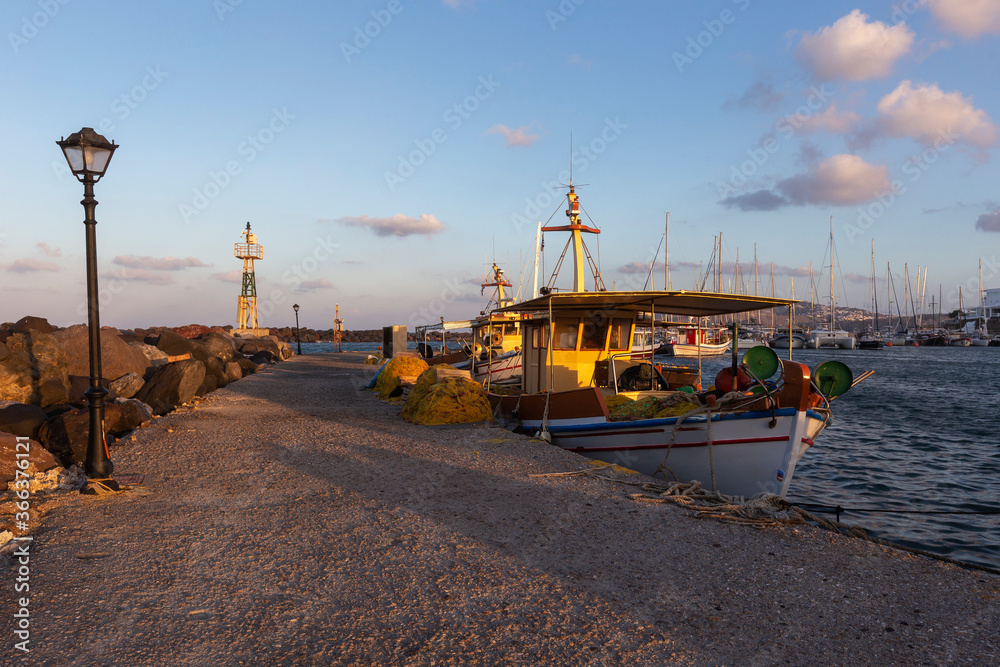 Naklejka premium Santorini, Marina Exomitis, a fishing boat moored at a stone pier in the port of Vlychada illuminated by the morning rays of the sun. There is a lamp on the pier and a metal lighthouse in the backgrou