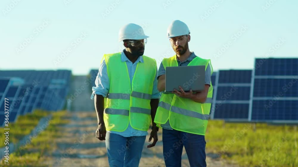 Two engineers with laptop on the solar farm in special uniform standing discuss the installation of sunny batteries electricity african american environmental slow motion Two engineers with laptop on the solar farm in special uniform standing discuss the installation of sunny batteries electricity african american environmental slow motion