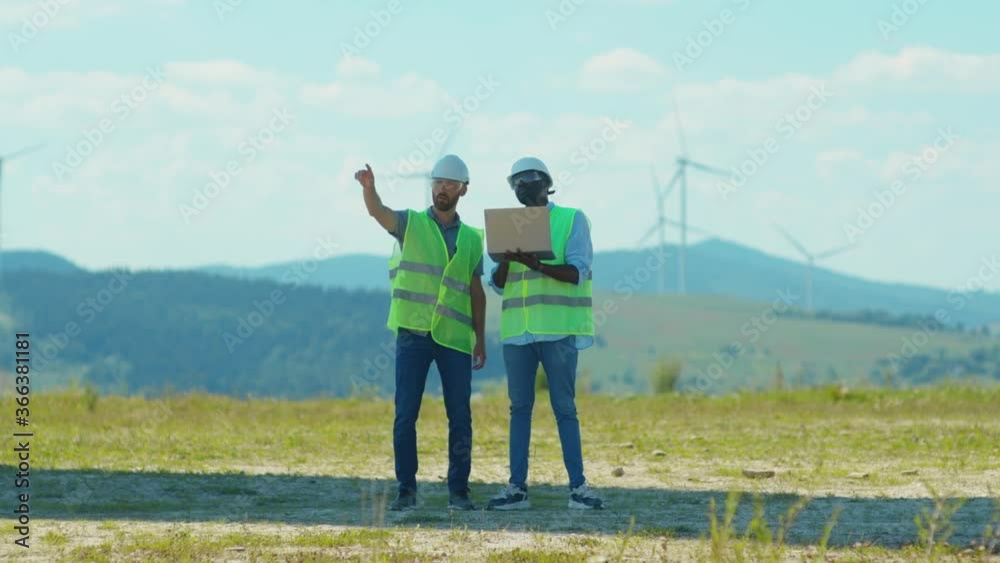 At sunny day two young engineers use laptop stand near windmills in fields team work discuss labor collaboration contribution windpower farm technicians talking close up slow motion At sunny day two young engineers use laptop stand near windmills in fields team work discuss labor collaboration contribution windpower farm technicians talking close up slow motion
