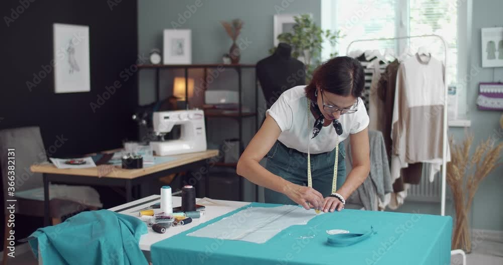Serious woman with brown hair working on design of new blue dress for client. Female designer using pattern and fabric at modern studio Serious woman with brown hair working on design of new blue dress for client. Female designer using pattern and fabric at modern studio