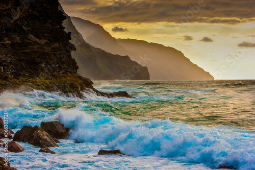 Waves crash at sunset on the rocks with the Na Pali headlandsin the background on the north shore of Kauai, Hawaii.