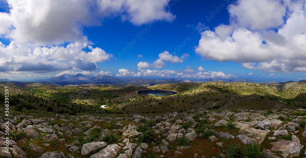 Wide panoramic view of the island of Crete in Greece. Landscape with a view of a water quarry, mountains and blue sky in clouds, with rocks and grass bushes in the foreground