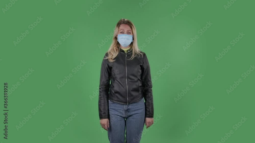 Portrait of beautiful young woman in medical protective face mask looking at camera worries and scared. Blonde with loose hair in a black jacket and jeans on a green screen in the studio. Health Portrait of beautiful young woman in medical protective face mask looking at camera worries and scared. Blonde with loose hair in a black jacket and jeans on a green screen in the studio. Health