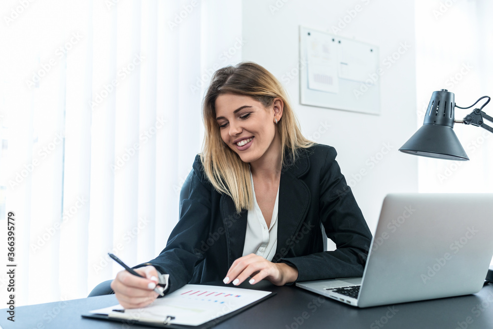 Smiling female entrepreneur in suit sitting in office and writing on paper. Smiling female entrepreneur in suit sitting in office and writing on paper.
