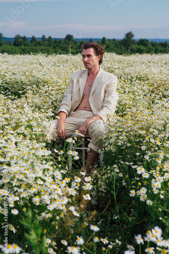 Tall handsome man sitting on a white chair in camomile flowers field © Smile