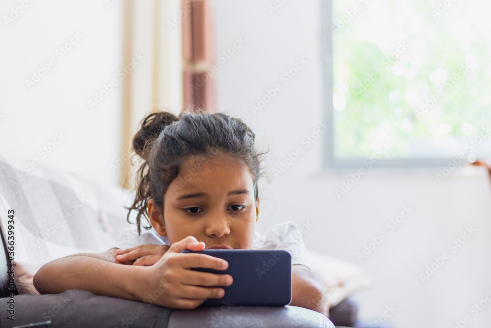 Brazilian kid Using Mobile Phone Watching Cartoons At Home, Lying On Sofa Relaxing On Weekend. Kid And Gadget Concept Brazilian kid Using Mobile Phone Watching Cartoons At Home, Lying On Sofa Relaxing On Weekend. Kid And Gadget Concept