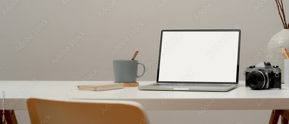 Blank screen laptop on worktable with camera, supplies, cup, decoration and office chair in home office Blank screen laptop on worktable with camera, supplies, cup, decoration and office chair in home office