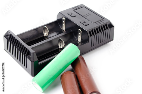 A black charger for two 18650 batteries. A green and brown 18650 battery on a white background next to the discharge, on charging. Male hand puts the battery on charge