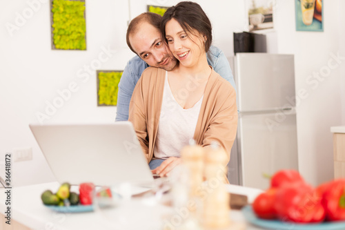 Smiling couple using laptop in kitchen with healthy vegetables on the table. Happy loving cheerful romantic in love couple at home using modern wifi wireless internet technology