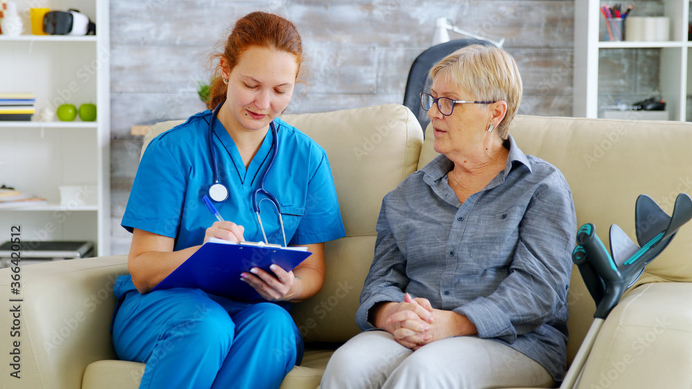 Female doctor with red hair taking notes on clipboard while talking with senior woman on the couch in nursing home Female doctor with red hair taking notes on clipboard while talking with senior woman on the couch in nursing home