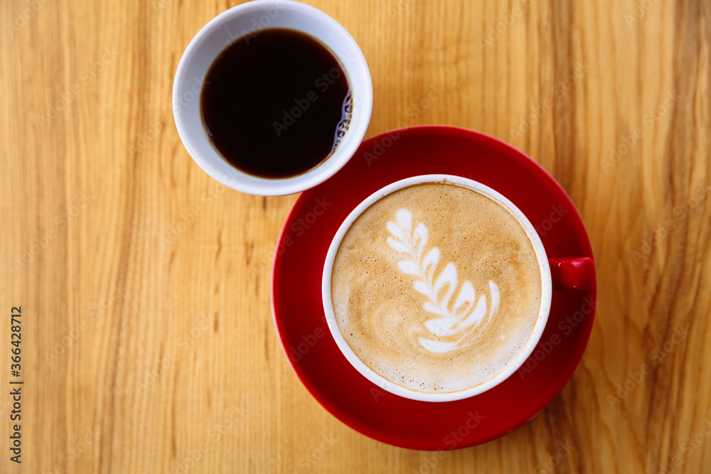 Aromatic coffee in a red cup with milk foam and latte art and freshly brewed coffee in a white cup on a light wooden table. Flat lay Aromatic coffee in a red cup with milk foam and latte art and freshly brewed coffee in a white cup on a light wooden table. Flat lay