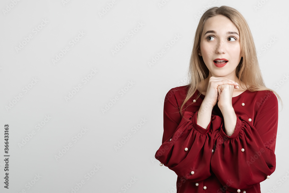 Photo of charismatic Pensive European young woman with long blonde hair keeps hands together near chin, has cute expression, wears red blouse. Copy space for your text Photo of charismatic Pensive European young woman with long blonde hair keeps hands together near chin, has cute expression, wears red blouse. Copy space for your text