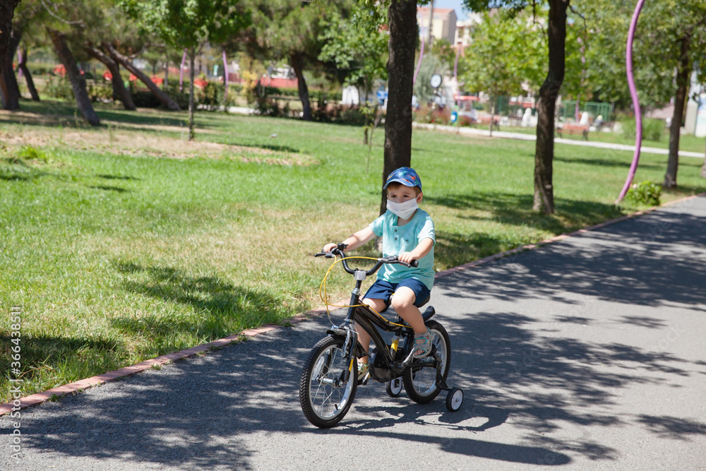Little cute boy with facial medical mask riding a bicycle in the park during beautiful sunny day, wear mask concept during new normal lifestyle. Healthcare and social distance concept.