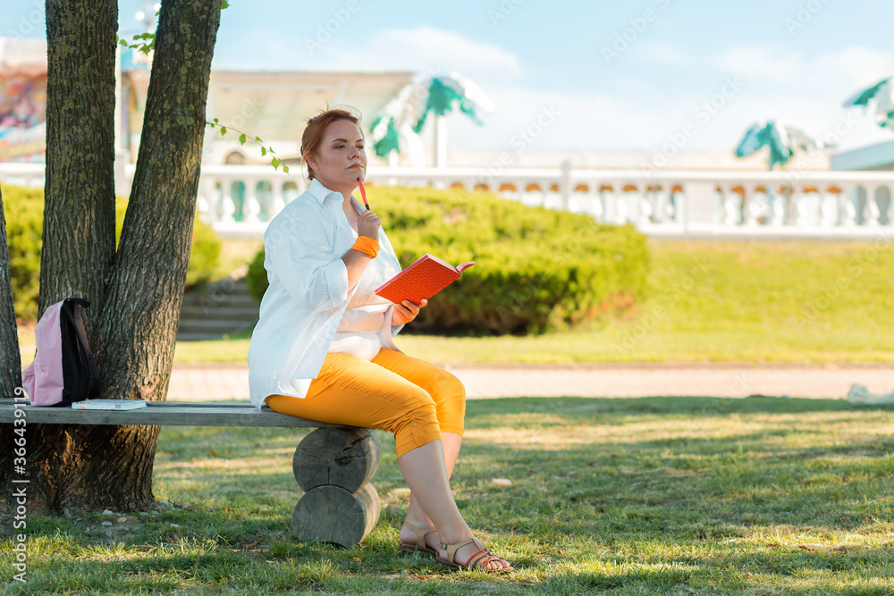 Fototapeta premium Education and leisure. Portrait of a plump woman sitting on a Park bench, looking thoughtfully, with a notebook and pen in her hands. Copy space