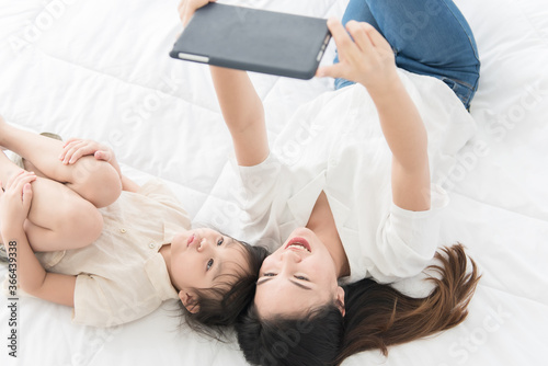 Top view Beautiful Asian mother and little daughter lying on a white bed in the house and smile and watching tablet together, feeling happy, family concept