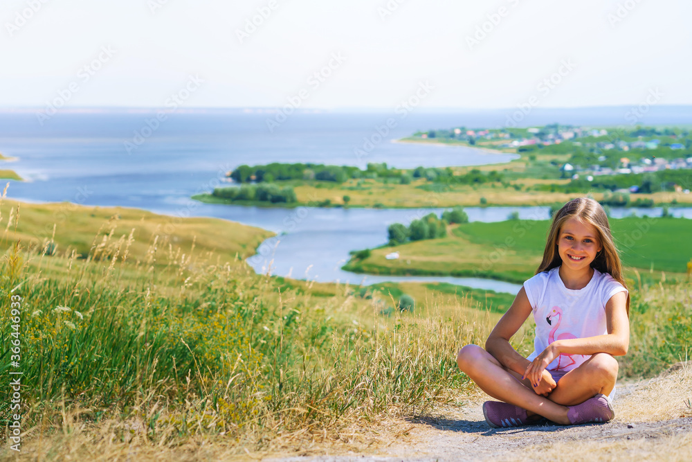 A little girl a child with long hair sits on a path with a mountain landscape with rivers and green meadows