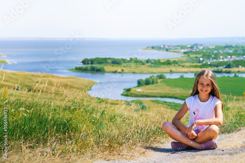 A little girl a child with long hair sits on a path with a mountain landscape with rivers and green meadows
