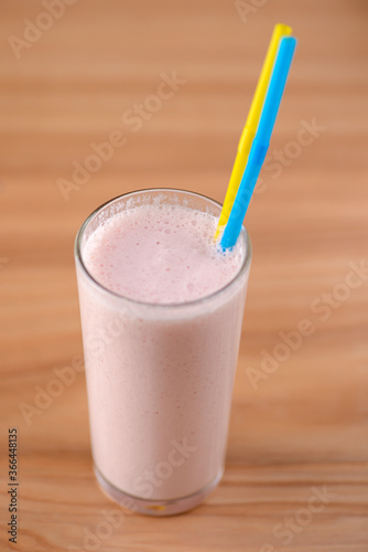 Vanilla strawberry milkshake in a tall galss with two plastic straws on light rustic wooden table.