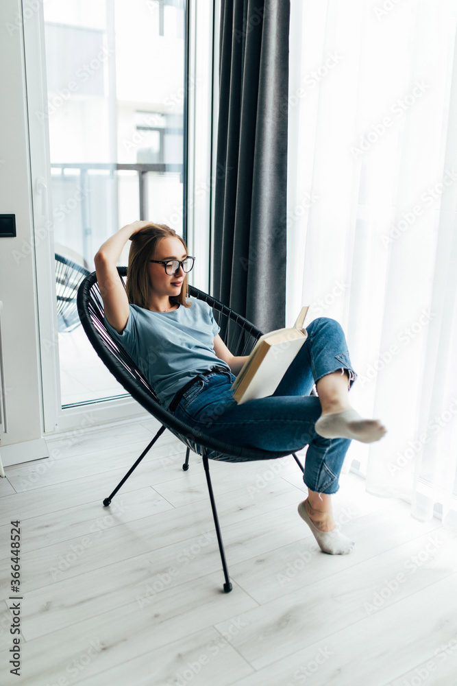 Young lazy woman at home sitting on modern chair in front of window relaxing in her living room reading book Young lazy woman at home sitting on modern chair in front of window relaxing in her living room reading book